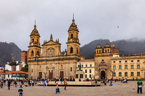 Bogota, Colombia - July 01, 2016: Looking from Plaza Bolivar, in the Andean Capital city of Bogota, in Colombia, South America, to the Eastern side of the Square. In full view is the Catedral Primada, the seat of the Roman Catholic Archbishop of Colombia. To the left of the image is the Casa del Florero and to the right, the Archbishop's chapel. and a section of his palace. The style of architecture is classical Spanish colonial. In the centre of the square is a statue of Simon Bolivar. In the far background in the haze of the overcast, cloudy sky, the Andes Mountains can be seen.; the peaks are obscured by clouds. People walk across the Square as they go about their daily work or explore the area. There are pigeons all over the square, and vendors sell grains to feed the pigeons. Photo shot in the afternoon sunlight, on a cloudy overcast day which works well to minimise harsh shadows; horizontal format. Copy space.