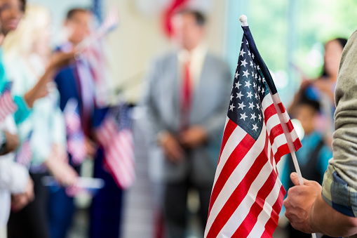 Focus on American flag in a diverse crowd of people. Supporters are cheering for a local candidate at a campaign rally or political town hall meeting.