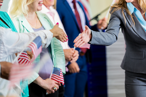 Unidentifiable female politician is shaking hands with supporters during a town hall meeting or political rally. Diverse people are lined up waving USA flags, and waiting to meet candidate.