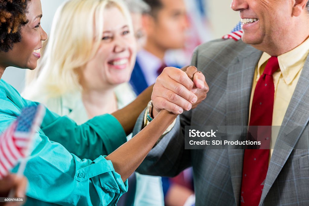 Candidate shaking supporter's hand during local political rally Mature adult Caucasian man is shaking hands with mid adult African American woman in crowd of cheering supporters. Diverse people are cheering for local candidate during political rally or town hall meeting. Candidate Stock Photo