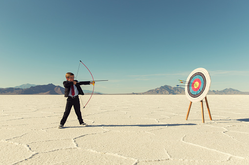 A young boy dressed in business suit, glasses and tie holds shoots a bow and arrow towards a target on the Bonneville Salt Flats in Utah. His business has found success and it mark for the future. He is smiling at the camera in front of blue skies.