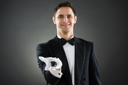 Portrait of happy young waiter giving car keys against gray background