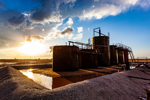 Farm oil storage tanks surrounded by water after rain with stormy sunset and sunflare. Metaphor: sun setting on oil production.  Horizontal, copy space, no people, names or logos.