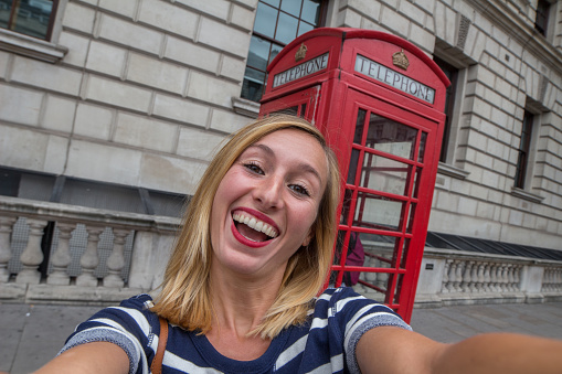 Young cheerful woman in London takes a selfie portrait with one of the classic London phone booth. Beautiful summer day.