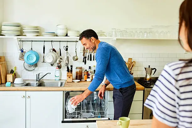 Mature man putting coffee mugs in dishwasher with his wife standing by