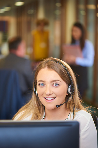 a young call centre representative greets a caller in a large open plan office . Co-workers can be seen defocussed in the background .