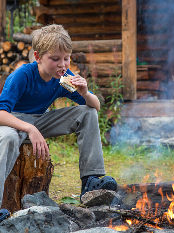 Boy eating marshmallow smore over campfire with rustic cabin in background