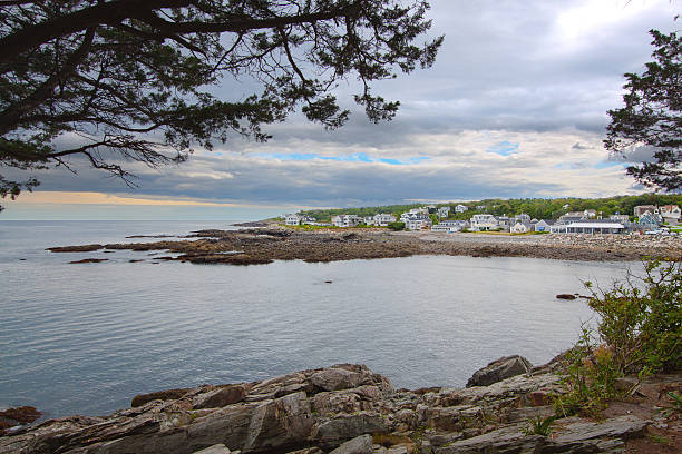 Harbor View with Rocks and Waterfront Houses, Perkins Cove, Ogunquit, Maine, USA. Ocean waters, rocky shore, green trees, bushes and blue sky with clouds are in the image. Canon EF 24-105mm f/4 L IS lens. HDR photorealistic image.