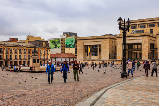 Bogota, Colombia - July 01, 2016: Looking towards the North Western corner of Bolivar Square in the Andean Capital city of Bogota in Colombia, South America. To the right of the image is the 'Palacio de Justicia' or translated, the Palace of Justice. It is the seat of the country's Supreme Court. To the left, is the Mayor's office and in the middle of the square, a statue of Simon Bolivar. Like many other squares across the world, there are plenty of pigeons on the square, waiting for visitors to feed them. The sky is overcast; it is drizzling; some people have their umbrellas open. Photo shot in the afternoon sunlight on a cloudy day; there are no harsh shadows. Horizontal format. Copy space.
