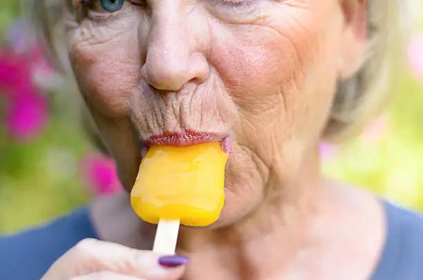 Senior woman sucking on an iced orange lolly outdoors on a summer day cooling off from the heat, close up cropped view on her mouth and lips