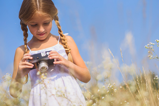 Cute female kid is holding camera and looking at shots with joy. She is standing on field and gently smiling