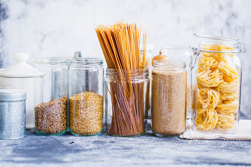 Couscous, linen seeds, noodles, quinoa, linguini pasta in glass jars over grey kitchen table. 