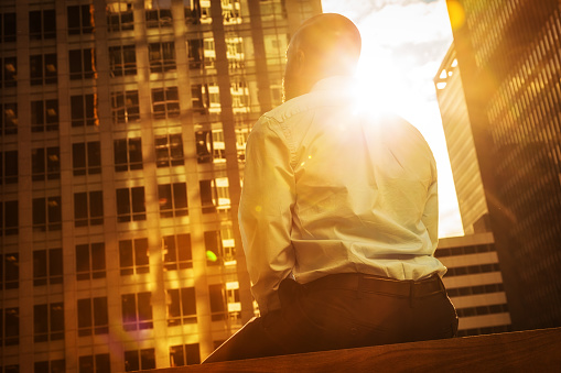 Rear-view photo of an African American businessman looking upward and contemplatively toward a city's skyline bathed in golden sunlight.