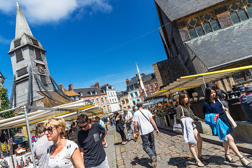 Honfleur, France - July 25, 2016: Tourists and locals strolling in the summer sun outside Saint Catherine's Church on Place Sainte-Catherine in the town of Honfleur in the Normandy region of France. The town was a favourite of many artists, and of the musician Erik Satie, and nowadays enjoys a thriving summer tourist industry, attracted by the culture and its open air markets. The building on the left is the wooden bell tower of Sainte Catherine's Church opposite.