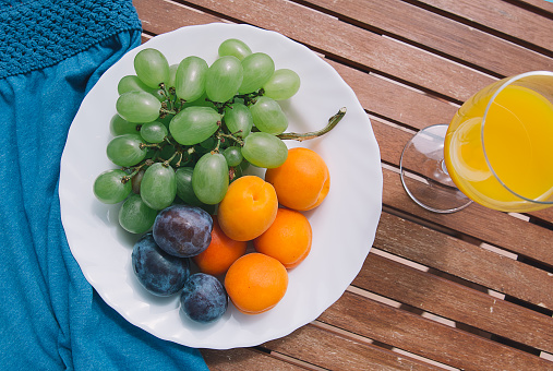 Summer lunch break or breakfast. Plate with fresh mixed fruits on the wooden table with a glass of juice and summer clothes. Assortment of juicy fruits.