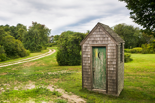 A small rustic privvy in a field next to an unpaved road in New England.