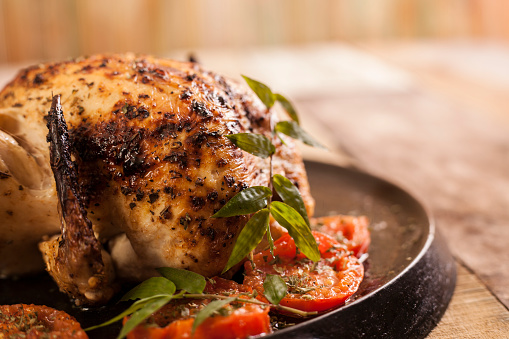Roasted whole chicken or small turkey with tomatoes in a cast iron skillet on rustic wooden table.  Healthy eating.  No people