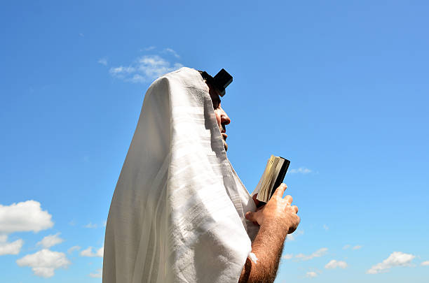Rear view color image depicting a Jewish man in his 30s, wearing a skull cap embroidered with the Star of David, praying alone inside the synagogue. Focus on the man with the place of worship defocused beyond. Room for copy space.