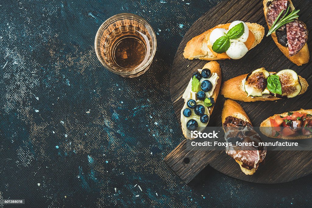 Italian crostini and glass of rose wine over dark background Italian crostini with various toppings on round wooden board and glass of rose wine over black plywood background, top view, copy space, horizontal composition Fig Stock Photo