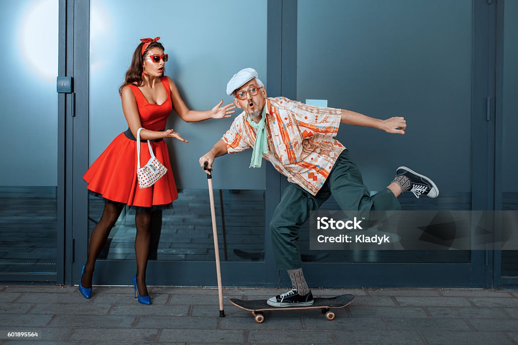 Funny old man riding a skateboard. Funny old man riding a skateboard, standing next to in surprise woman. Senior Men Stock Photo