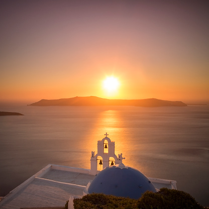 From high ground view point, a majestic sunset takes place in the horizon of the Santorini volcanic caldera. The blue dome accompanied with a bell tower, which is the symbol of the Cyclades islands in Greece, sits right on the brightly sun lit surface of the sea.