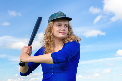 Young dutch woman with baseball bat and cap against blue sky. The dutch teenage girl is 18 years old and she looks rather sturdy and tough on this photo. I took the image on a sunny day in summer to get a background with blue sky. Concept of active,activity,sport,sports,baseball,bat,cap,long hair,summer,summertime,ready,hit,play,game.