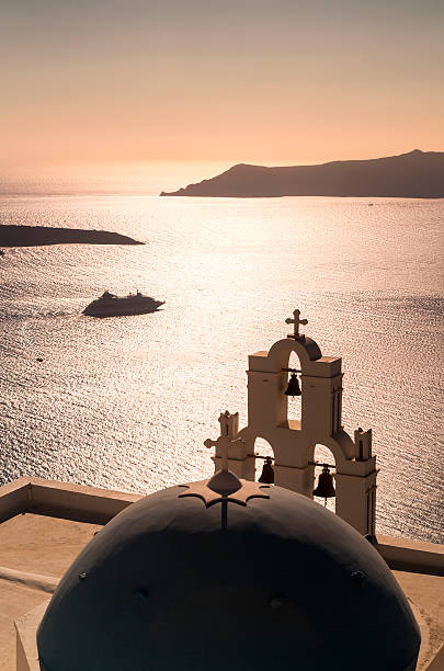 From a high ground view point, a sunset takes place over the horizon of the worldwide famous island of Santorini of Greece. In the volcanic caldera far below, a cruise ship carrying visitors is cruising to another destination. On the foreground, the dome accompanied with a bell tower is the Orthodox symbol of the Cyclades islands and stands right on the brightly sun lit surface of the sea.