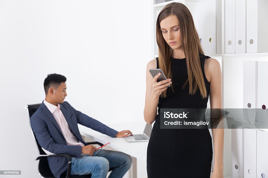 It's brave new world Businesswoman in black elegant dress is checking her phone. Her Asian colleague is working with his laptop Adult Stock Photo