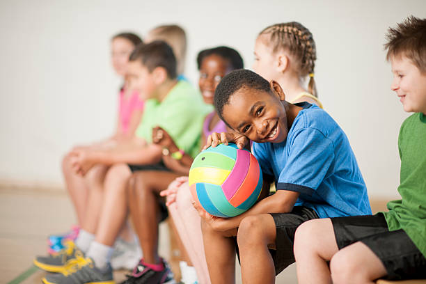 Sitting on the Bench Before the Game A multi-ethnic group of elementary age children are sitting in gym class and are going to play a game of volleyball. One boy is smiling while looking at the camera. young volleyball girl stock pictures, royalty-free photos & images
