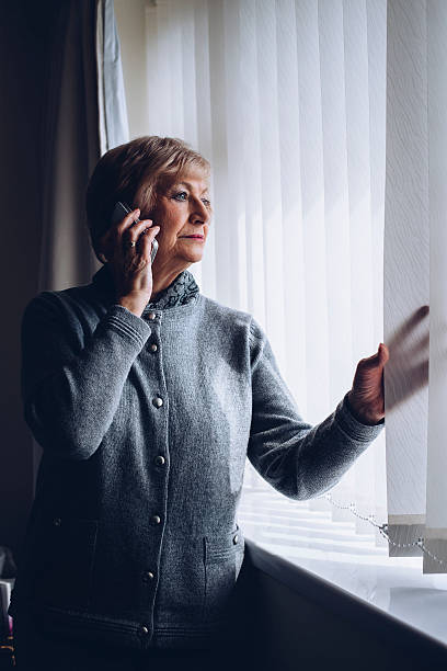 Reporting a Crime Senior woman talking on the phone whilst looking out of her window. neighborhood crime watch stock pictures, royalty-free photos & images