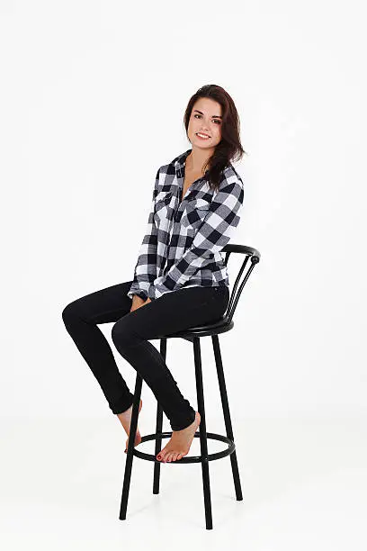 Awesome caucasian attractive sexy professional female model with brown hair posing in studio wearing casual shirt and black ragged jeans, sitting on chair, smiling, isolated on white background