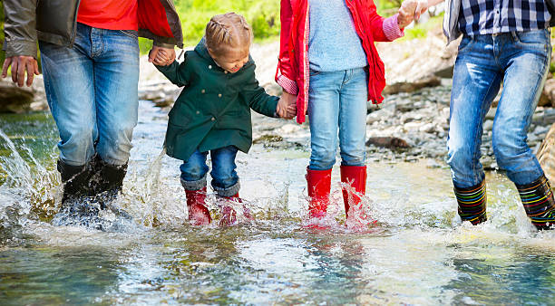 glückliche familie trägt regenstiefel, die in einen bergfluss springen - father directly below child looking up stock-fotos und bilder