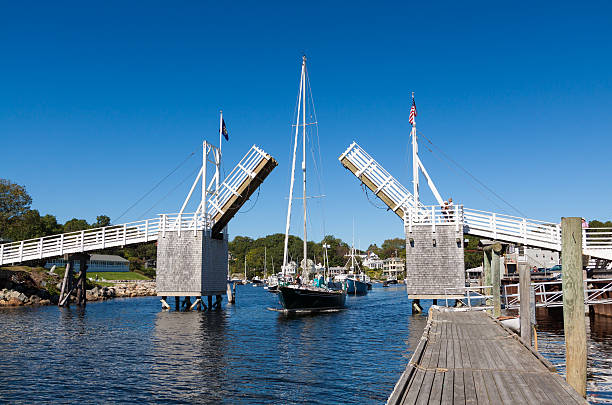 Harbor View with Sailboat Passing Under Drawbridge, Perkins Cove, Maine. Ogunquit, ME, USA - September 18, 2013: Harbor View with Sailboat Passing Under Drawbridge lit by the morning sun, Perkins Cove, Ogunquit, Maine, New England. Sailors steering the boat, sightseeing tourists, water of the cove, footbridge, pier, american flag and vivid blue sky with clouds are in the image. Polarizing filter. sailboat mast stock pictures, royalty-free photos & images