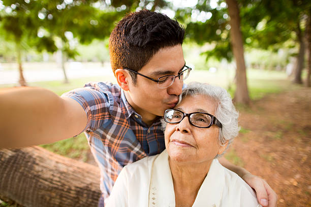 Selfie of grandson kissing grandmother on forehead A selfie of a latin adult grandson kissing his senior grandmother on the forehead in a horizontal waist up shot outdoors. hispanic grandmother stock pictures, royalty-free photos & images