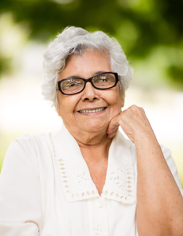 A portrait of a senior latin woman smiling at the camera with hand on chin in a vertical head and shoulder shot.