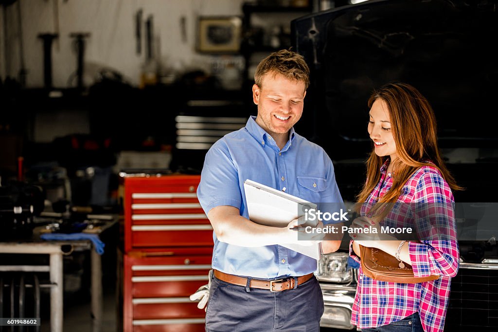 Happy customer discusses repairs with auto mechanic in repair shop. Happy customer!  A smiling Latin descent woman discusses automobile repair invoice with an auto mechanic in a repair shop.  She is discussing the vehicle's repairs with the mechanic, who is explaining her service.  Toolbox, workshop background. Unidentifiable, SUV-style vehicle. Auto Repair Shop Stock Photo