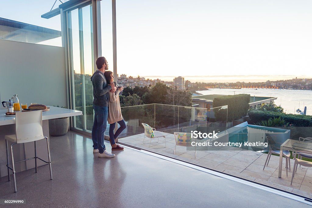 Couple having breakfast together at home. Couple having breakfast together at home. They are standing in the kitchen drinking coffee. The woman is happy and smiling. They are in love and very affectionate. The house is very luxurious and has a swimming pool and water view of the ocean. Wide angle Luxury Stock Photo