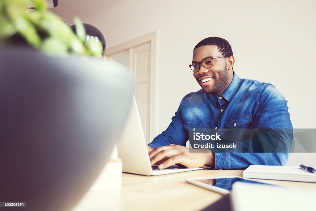 Afro american young man typing on laptop in an office Happy afro american young man wearing denim shirt using laptop at home or in the office.  Computer Stock Photo