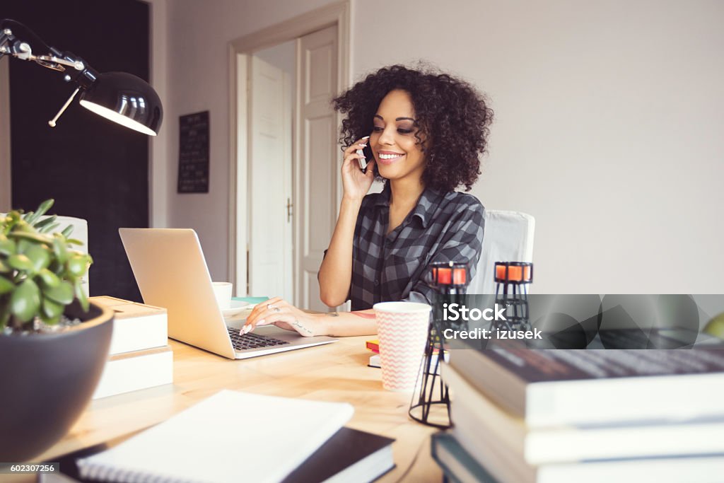 Afro young woman in the home office, using laptop Afro young woman sitting at the desk in a home office, using laptop and talking on mobile phone. Office Stock Photo