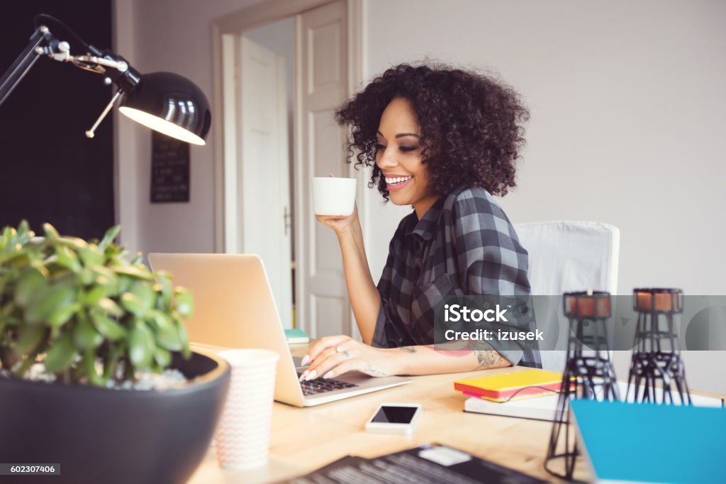 Afro young woman in the home office, using laptop Afro young woman sitting at the desk in a home office, using laptop, drinking coffee. Working At Home Stock Photo