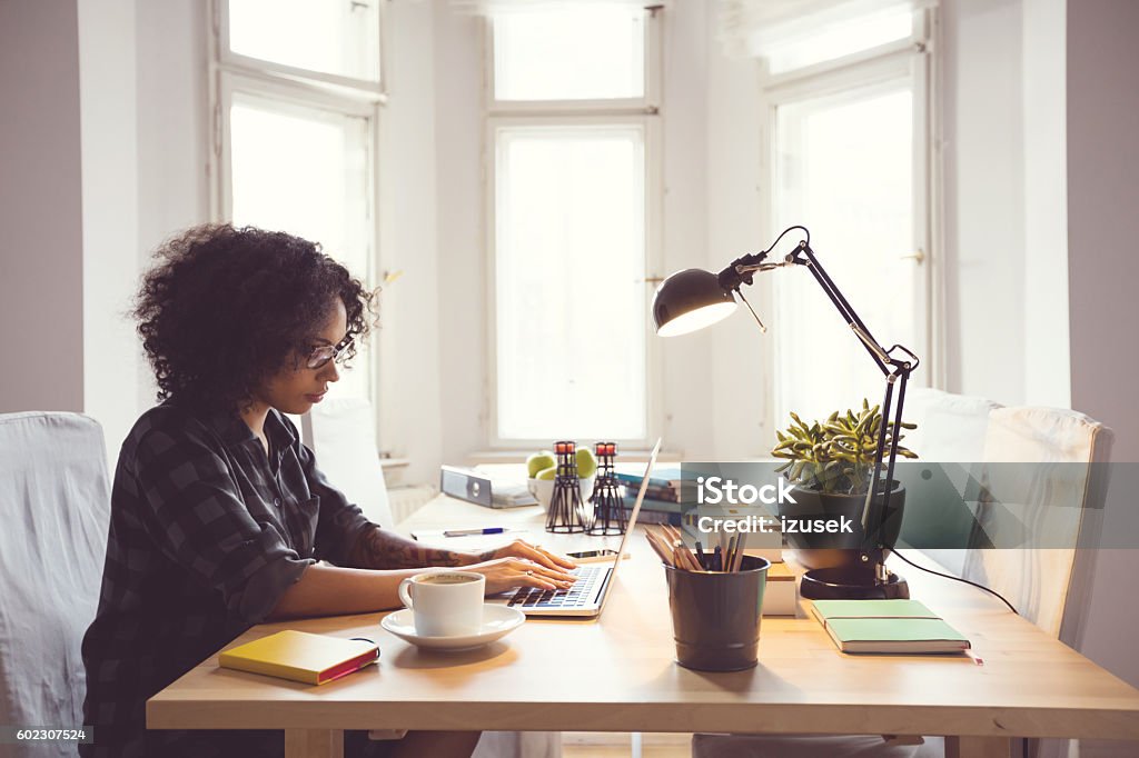 Afro young woman in the home office, using laptop Afro young woman sitting at the table in a home office, using laptop. Working At Home Stock Photo