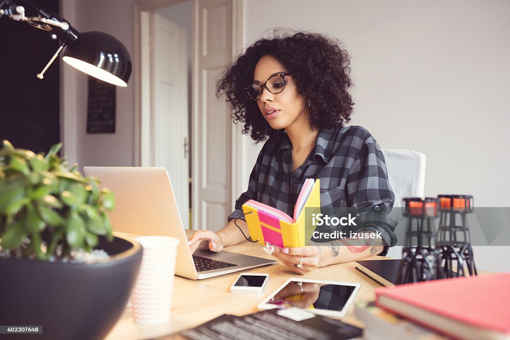 Afro young woman in the home office, using laptop Afro young woman sitting at the desk in a home office, using laptop. Freelance Work Stock Photo