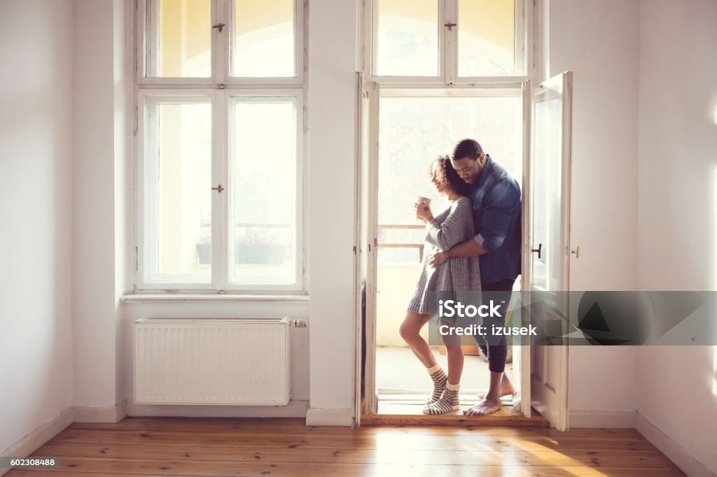 Afro american couple standing by the window at home Afro american couple embracing by the window at home in the morning light. Woman drinking coffee. Adult Stock Photo