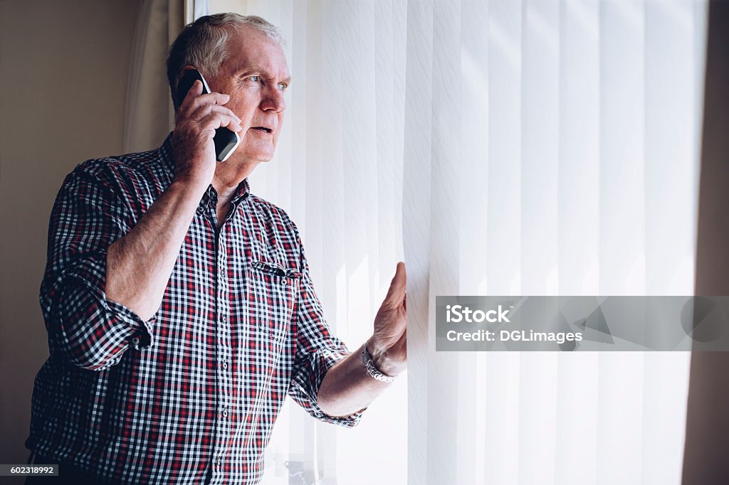 Reporting a Crime Senior man talking on the phone whilst looking out of his window. He has a worried look on his face. Using Phone Stock Photo