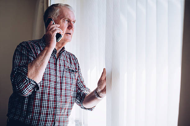 Reporting a Crime Senior man talking on the phone whilst looking out of his window. He has a worried look on his face. neighborhood crime watch stock pictures, royalty-free photos & images