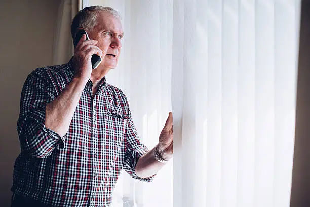 Senior man talking on the phone whilst looking out of his window. He has a worried look on his face.