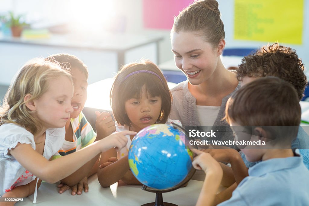 Teacher looking at schoolchildren touching globe Smiling female teacher looking at schoolchildren touching globe in classroom Preschool Stock Photo