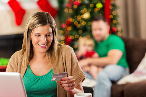 Pretty Caucasian woman doing online Christmas shopping on her laptop computer. Cheerful woman is sitting at a table looking down at her debit card, credit card to purchase Christmas presents online for her family.