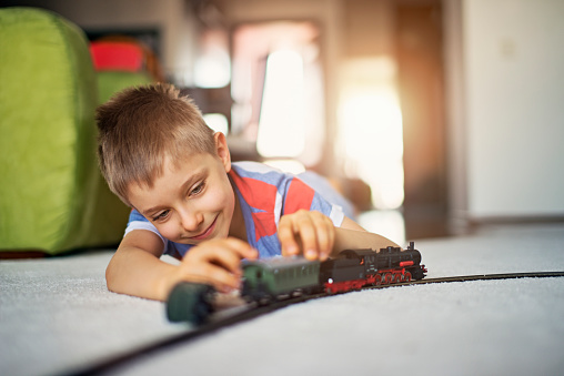 Happy little boy lying on a carpet, playing with miniature train. Very shallow depth of field. Sunny day indoors.