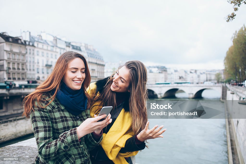 Two smiling women walking on bridge Smiling young women standing on Pont Notre dame and having fun. One wears checkered coat and blue scarf and holding a smart phone, other wears jacket and yellow scarf and looking at her. River and buildings on background. Adult Stock Photo